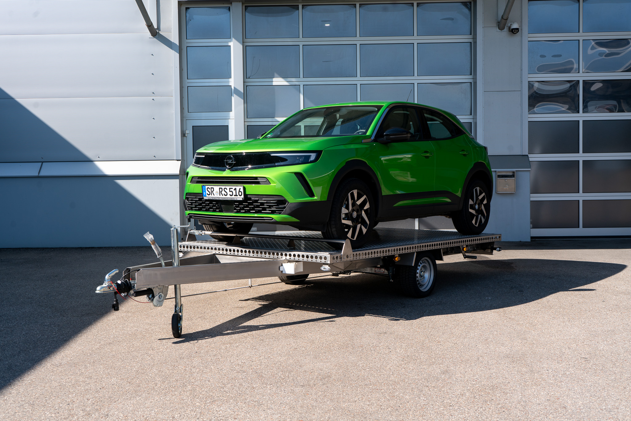 Edelstahl Plattformanhänger, mit einem grünen Opel Mokka auf dem Anhänger geladen - vor einem Hallentor - Frontansicht