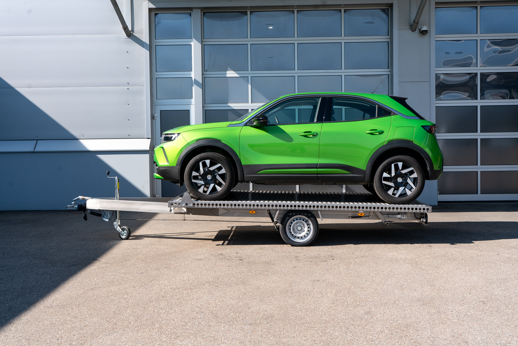 Edelstahl Plattformanhänger, mit einem grünen Opel Mokka auf dem Anhänger geladen - vor einem Hallentor - Seitenansicht