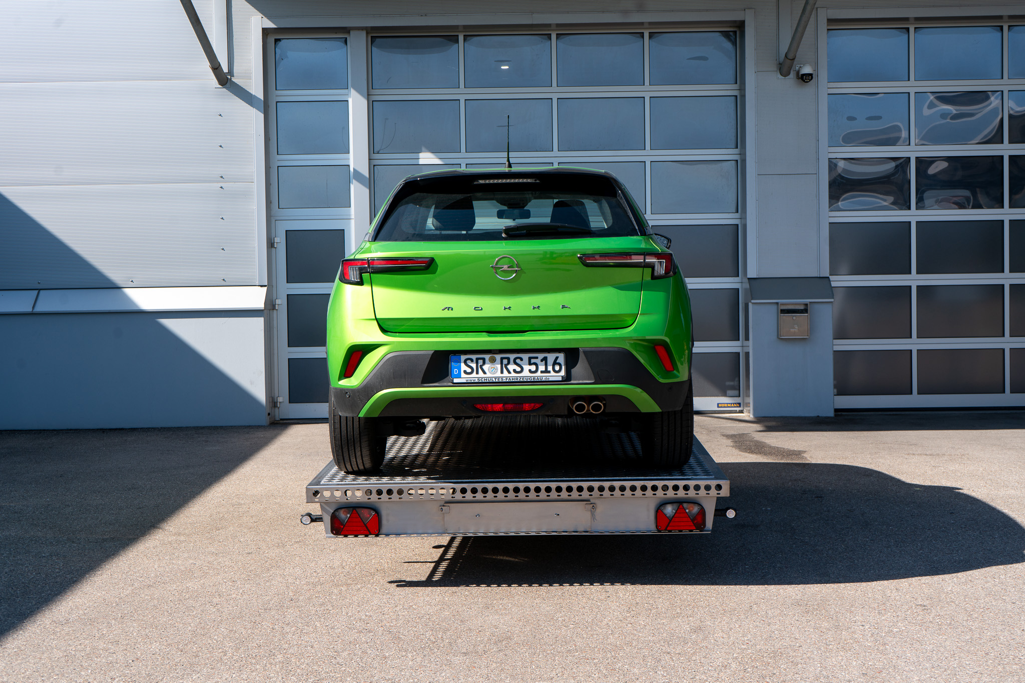 Edelstahl Plattformanhänger, mit einem grünen Opel Mokka auf dem Anhänger geladen - vor einem Hallentor - Heckansicht