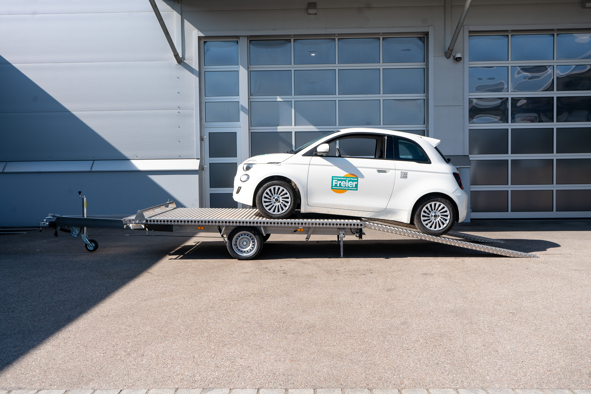 Edelstahl Plattformanhänger, mit einem weißen Fiat 500 auf dem Anhänger geladen - vor einem Hallentor - Seitenansicht