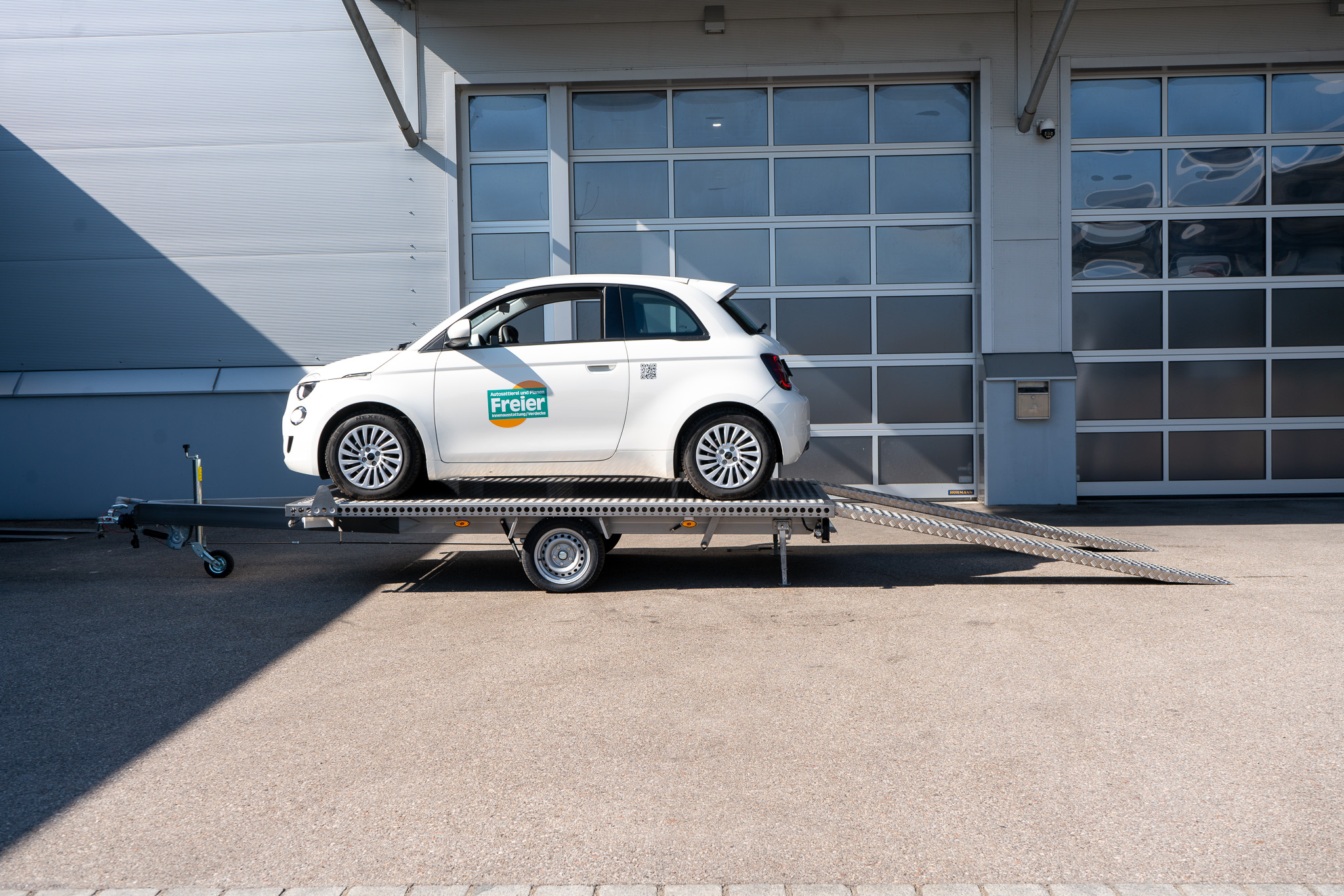 Edelstahl Plattformanhänger, mit einem weißen Fiat 500 auf dem Anhänger geladen - vor einem Hallentor - Seitenansicht