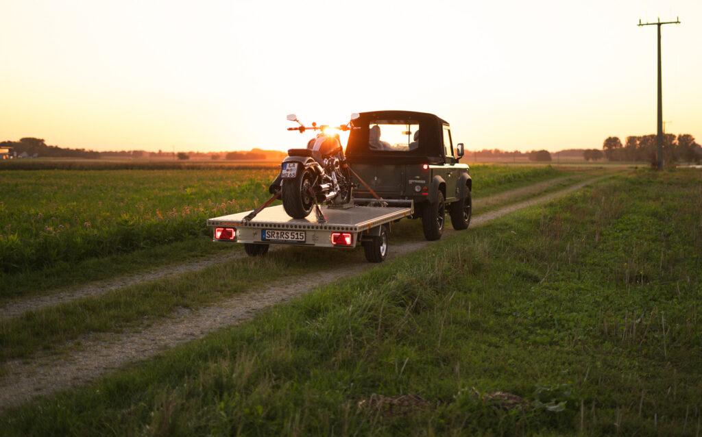 Aluminium Anhänger mit roten LED-Leuchten, beladen mit einer Harley Davidson, auf einem Feldweg bei Sonnenuntergang - Heckansicht