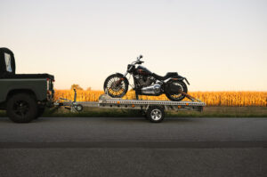 Aluminium Anhänger mit roten LED-Leuchten, beladen mit einer Harley Davidson, auf einem Feldweg bei Sonnenuntergang - Seitenansicht
