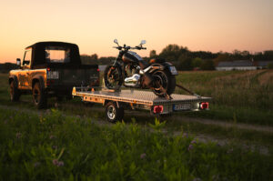 Aluminium Anhänger mit roten LED-Leuchten, beladen mit einer Harley Davidson, auf einem Feldweg bei Sonnenuntergang - Heckansicht