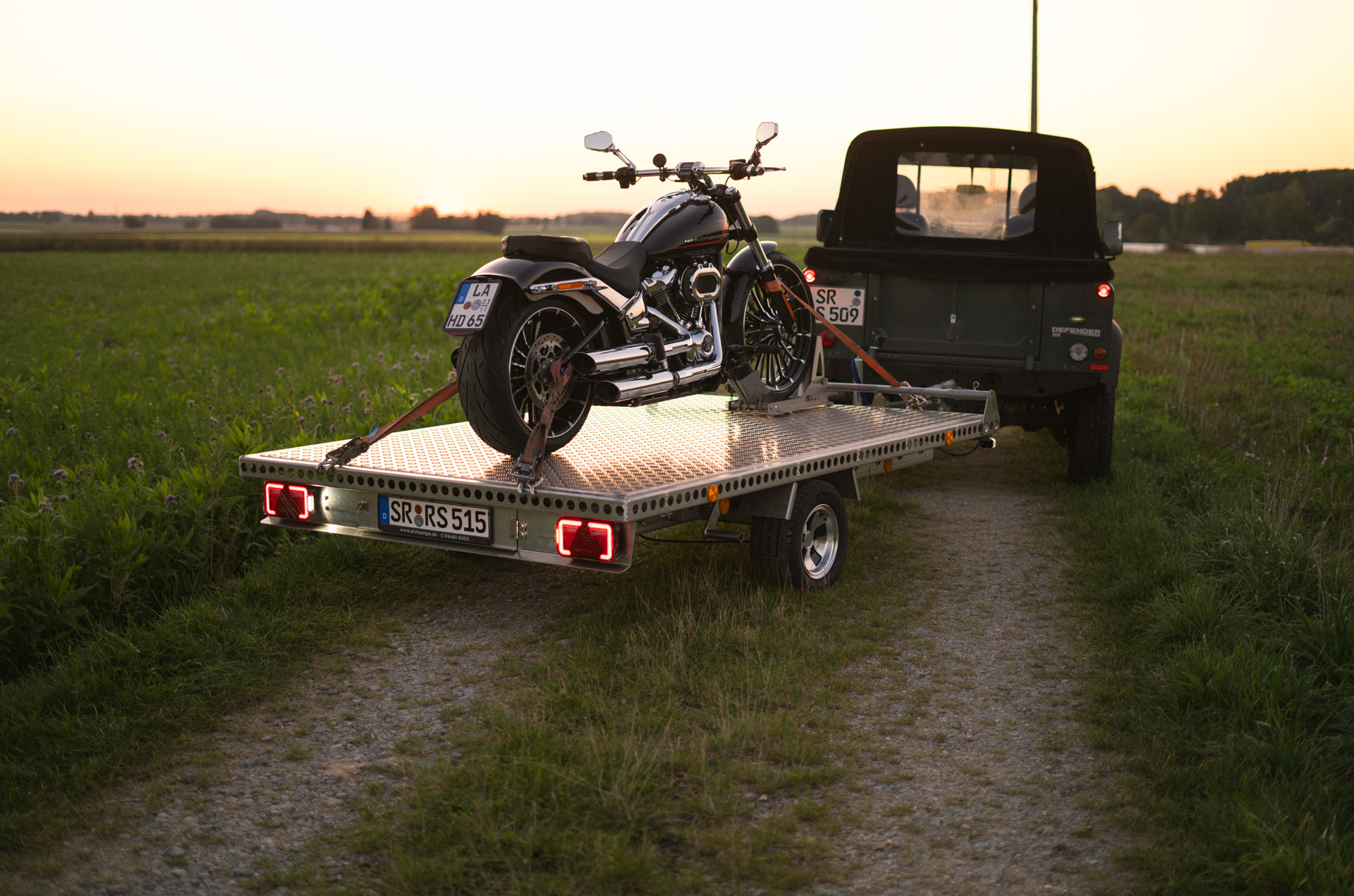 Aluminium Anhänger mit roten LED-Leuchten, beladen mit einer Harley Davidson, auf einem Feldweg bei Sonnenuntergang - Heckansicht