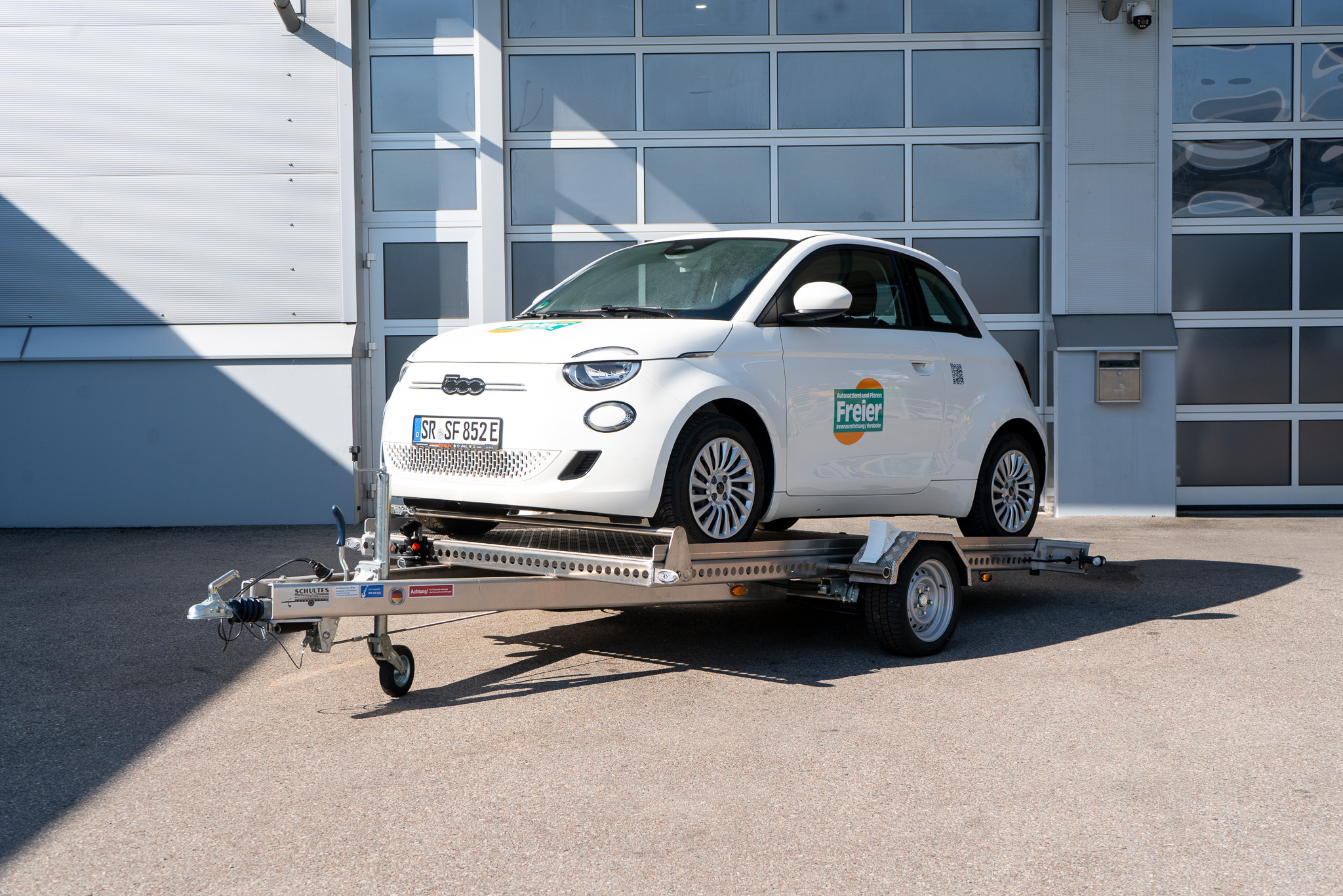 Absenkanhänger beladen mit weißem Fiat 500 auf dem Anhänger - Ansicht von vorne vor einem geschlossenen Hallentor