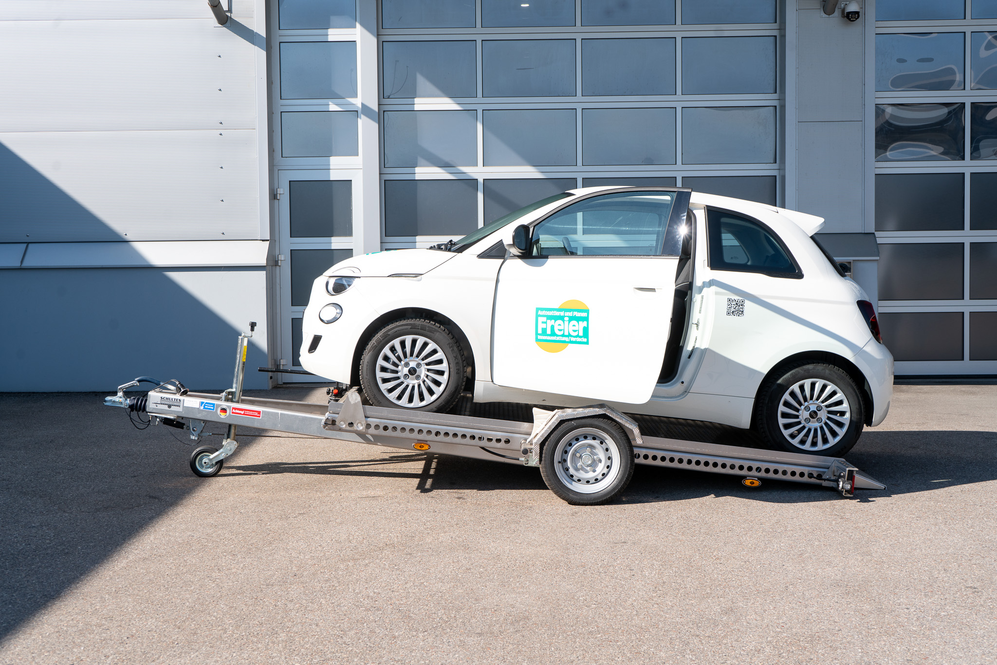 Absenkanhänger abgesenkt mit weißem Fiat 500 auf dem Anhänger - Ansicht von der Seite vor einem geschlossenen Hallentor