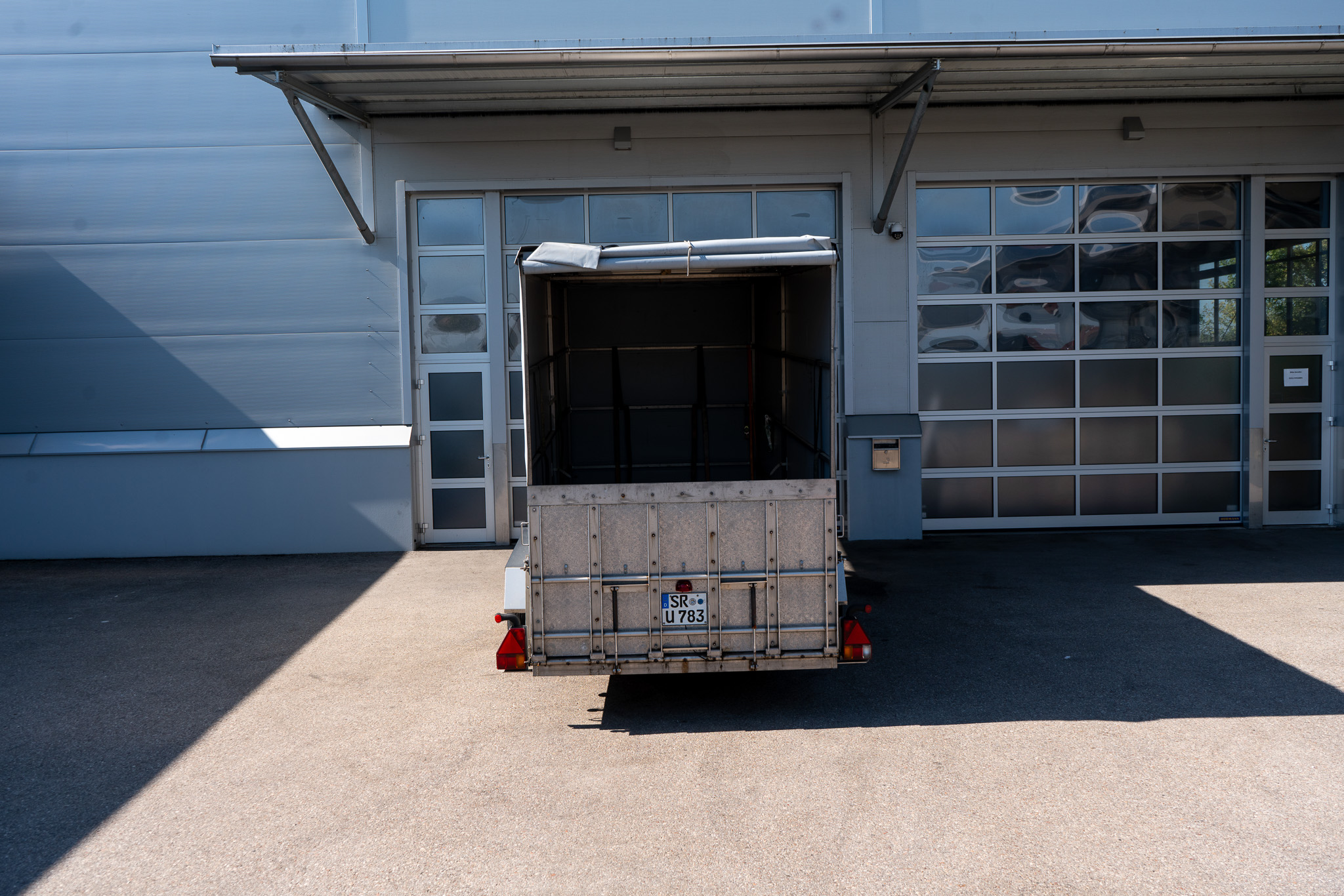 Absenkbarer Anhänger zum transport von Gabelstaplern mit grauer Plane vor einer Halle, von hinten