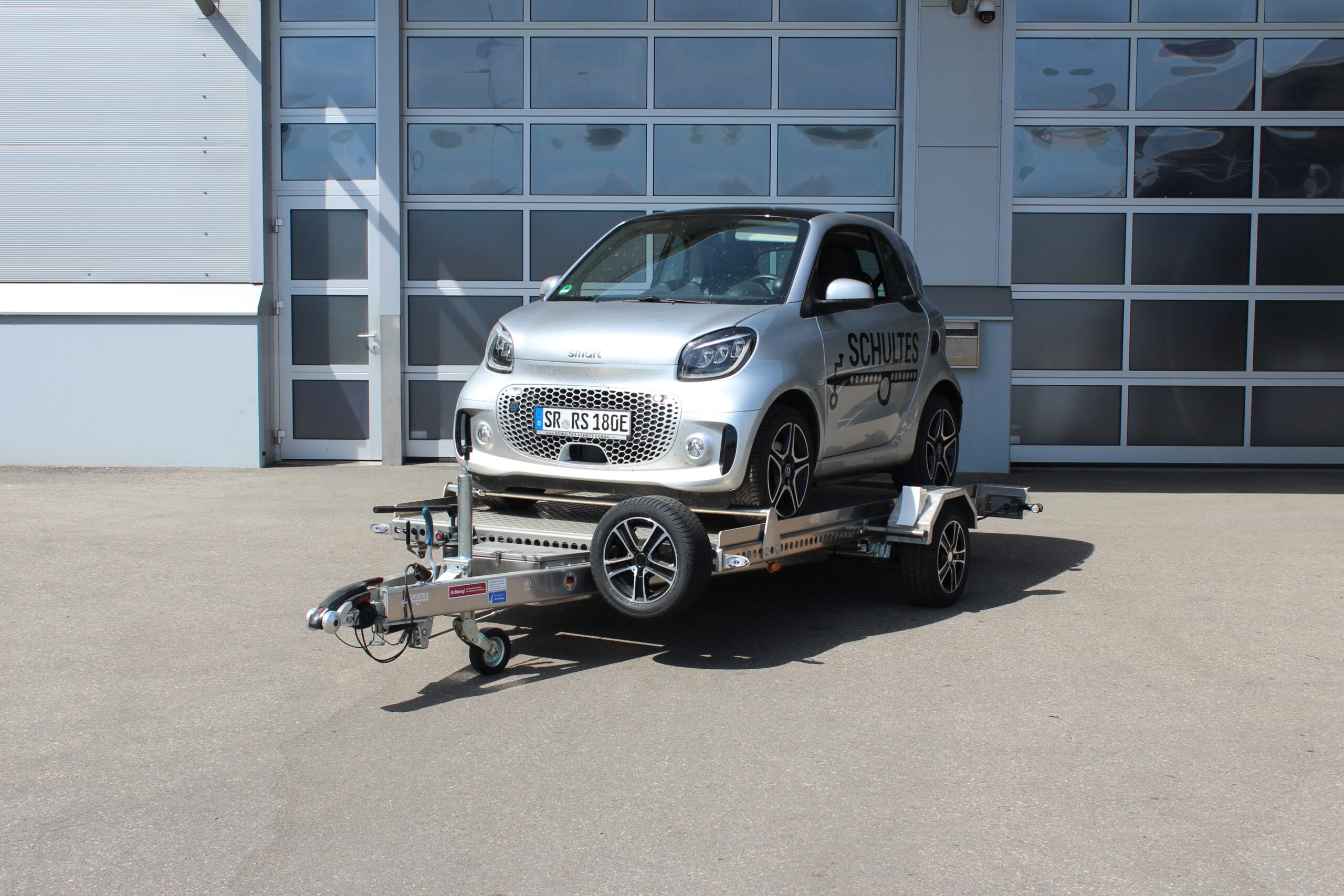Absenkanhänger aus Aluminium mit einem silbernen Smart fortwo beladen vor einer Halle in einem Hof