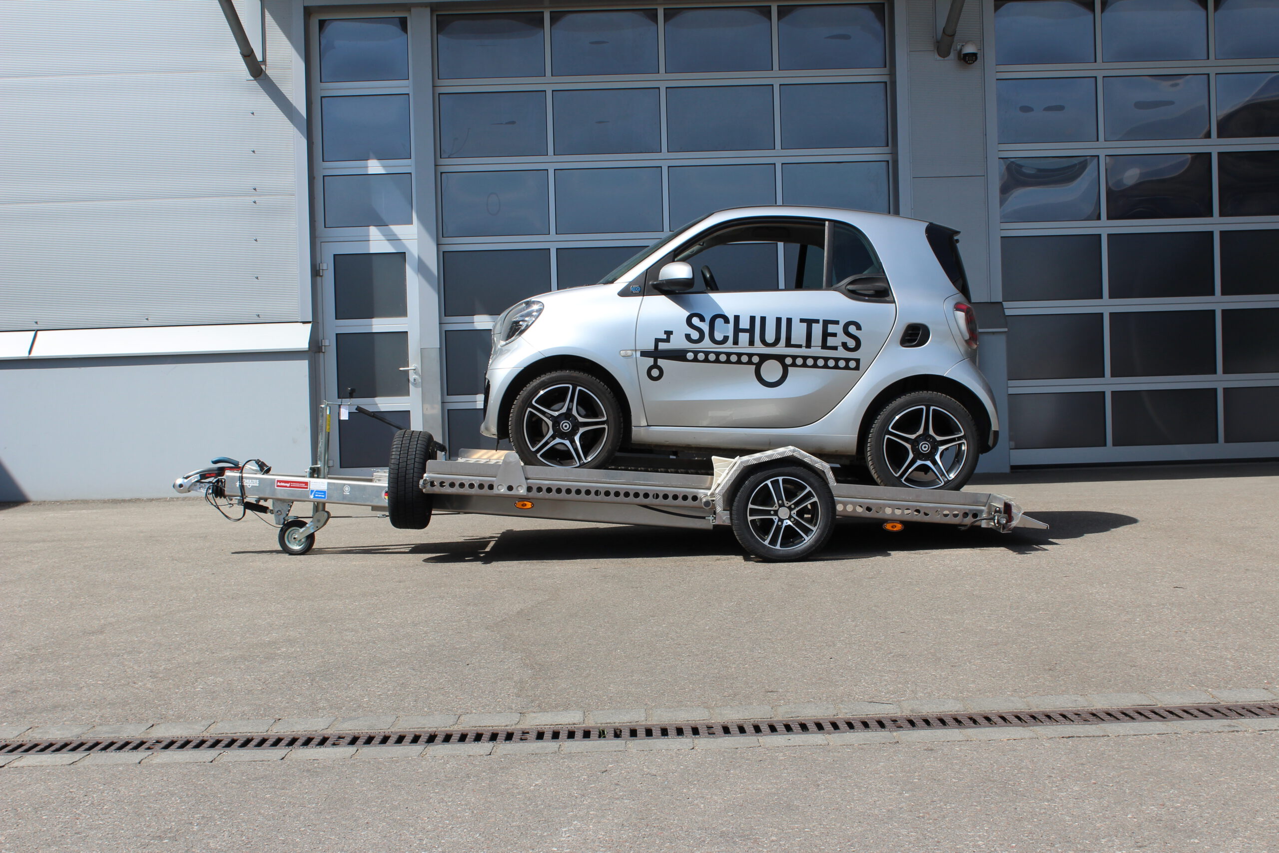Absenkanhänger aus Aluminium mit einem silbernen Smart fortwo beladen vor einer Halle in einem Hof