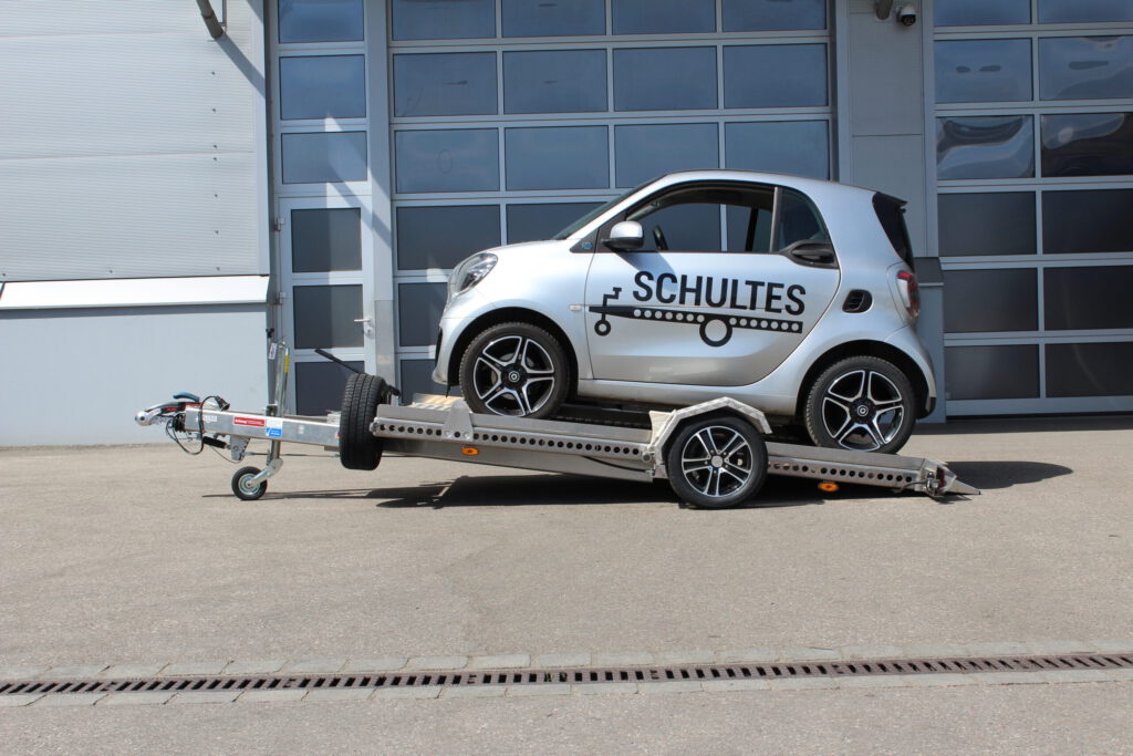 Absenkanhänger aus Aluminium mit einem silbernen Smart fortwo beladen vor einer Halle in einem Hof