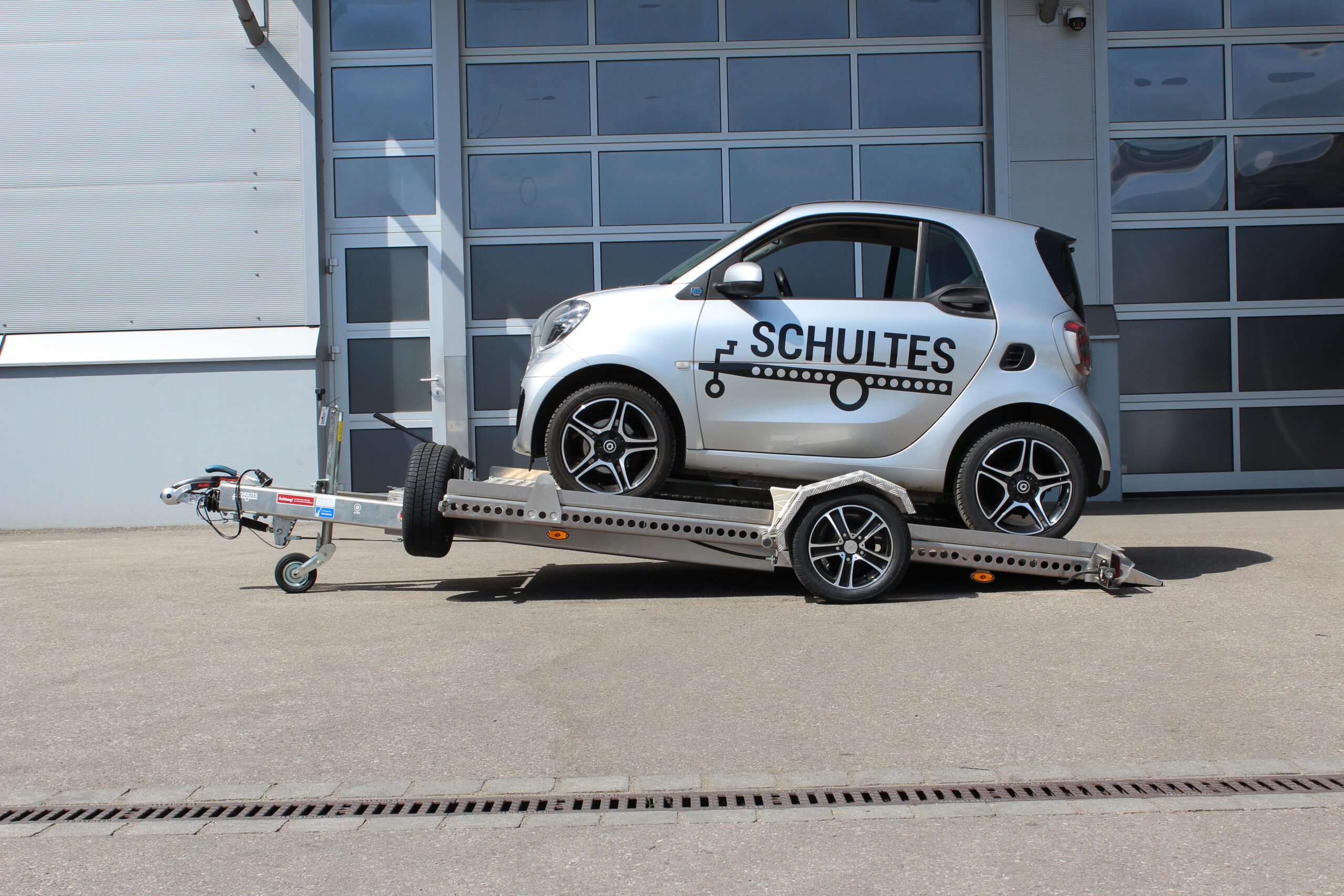 Absenkanhänger aus Aluminium mit einem silbernen Smart fortwo beladen vor einer Halle in einem Hof