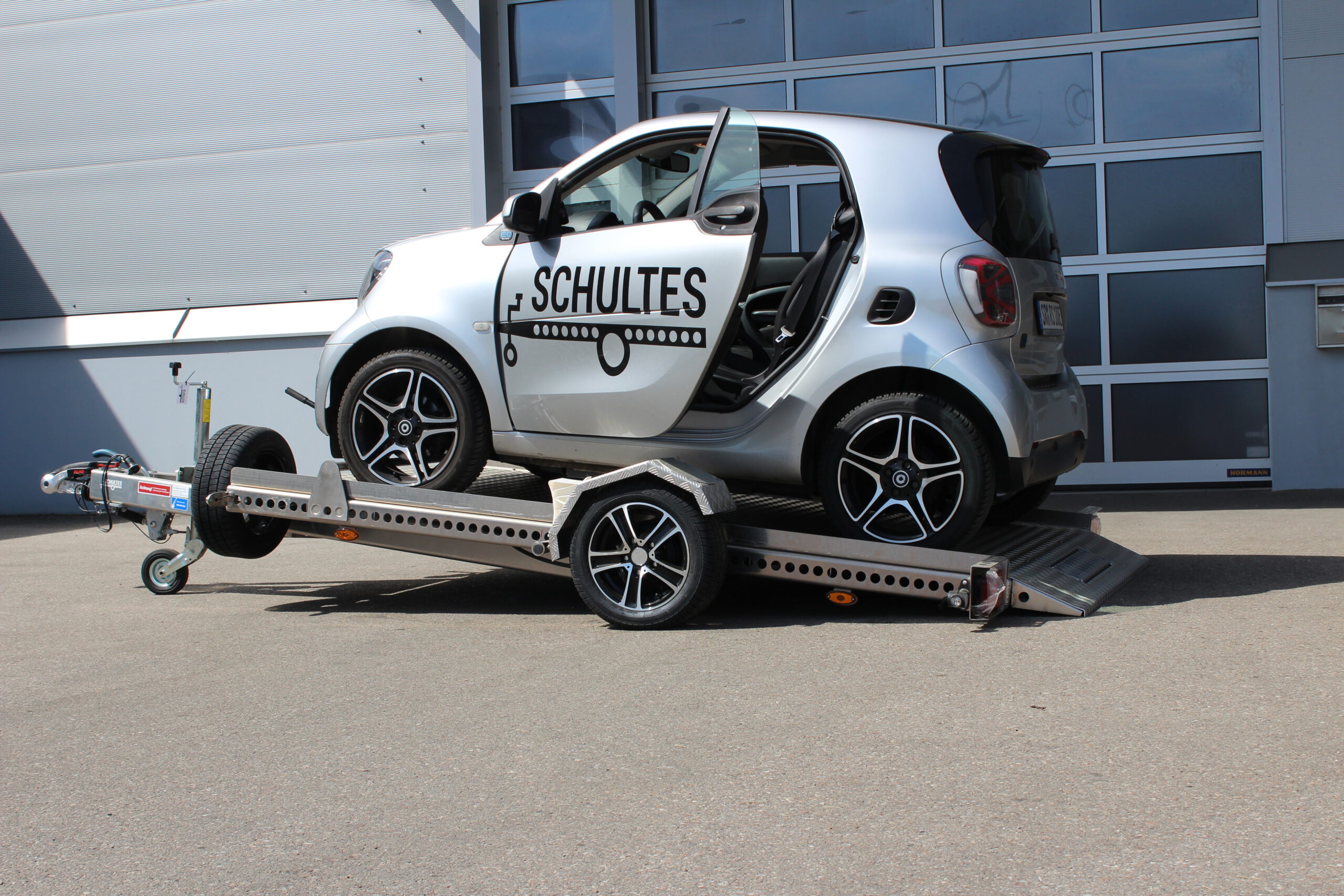 Absenkanhänger aus Aluminium mit einem silbernen Smart fortwo beladen vor einer Halle in einem Hof