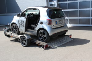 Absenkanhänger aus Aluminium mit einem silbernen Smart fortwo beladen vor einer Halle in einem Hof