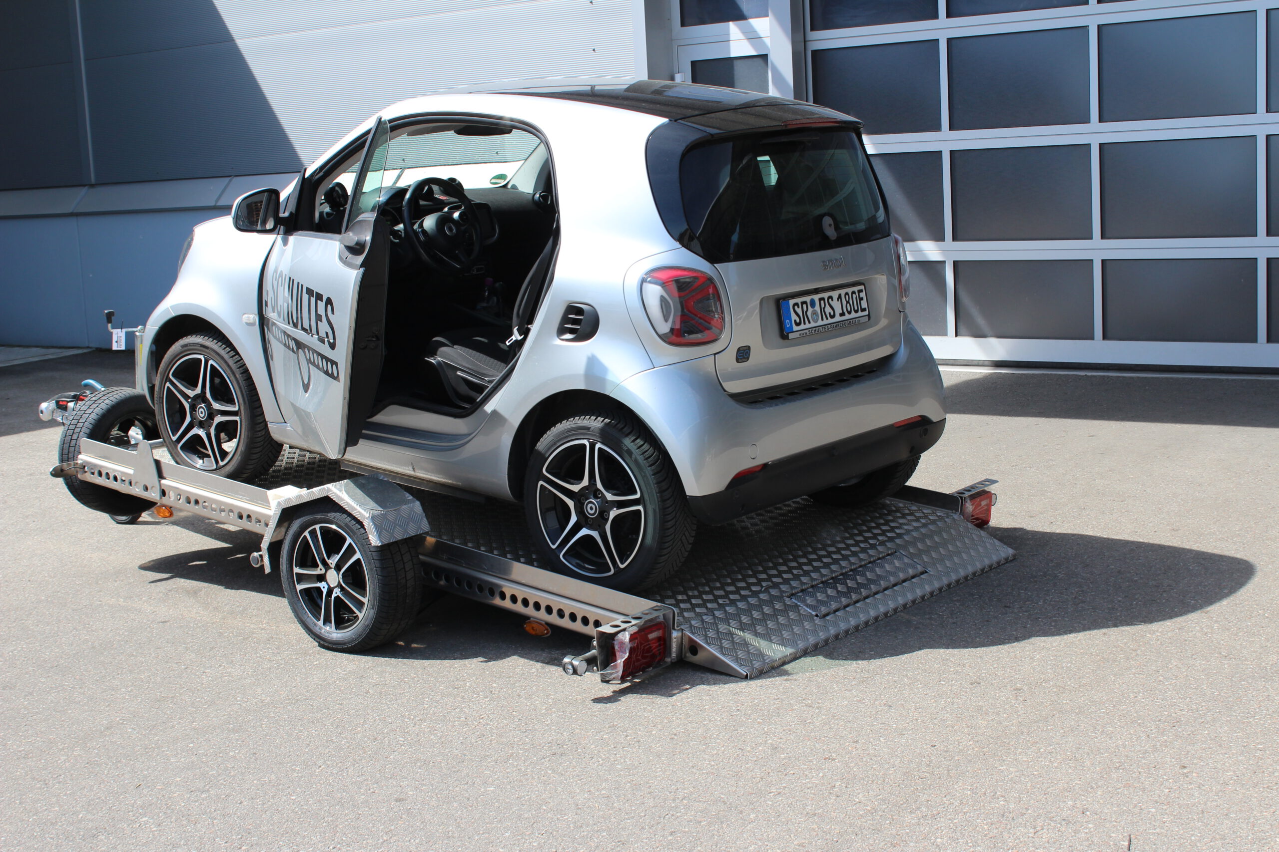 Absenkanhänger aus Aluminium mit einem silbernen Smart fortwo beladen vor einer Halle in einem Hof