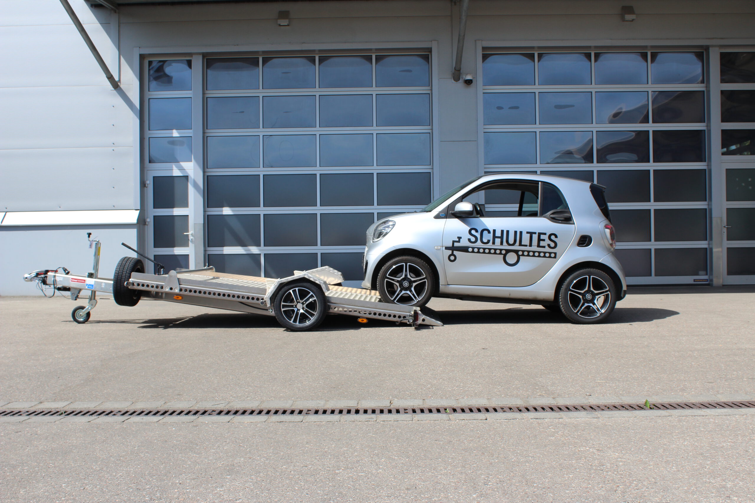 Absenkanhänger aus Aluminium mit einem silbernen Smart fortwo beladen vor einer Halle in einem Hof