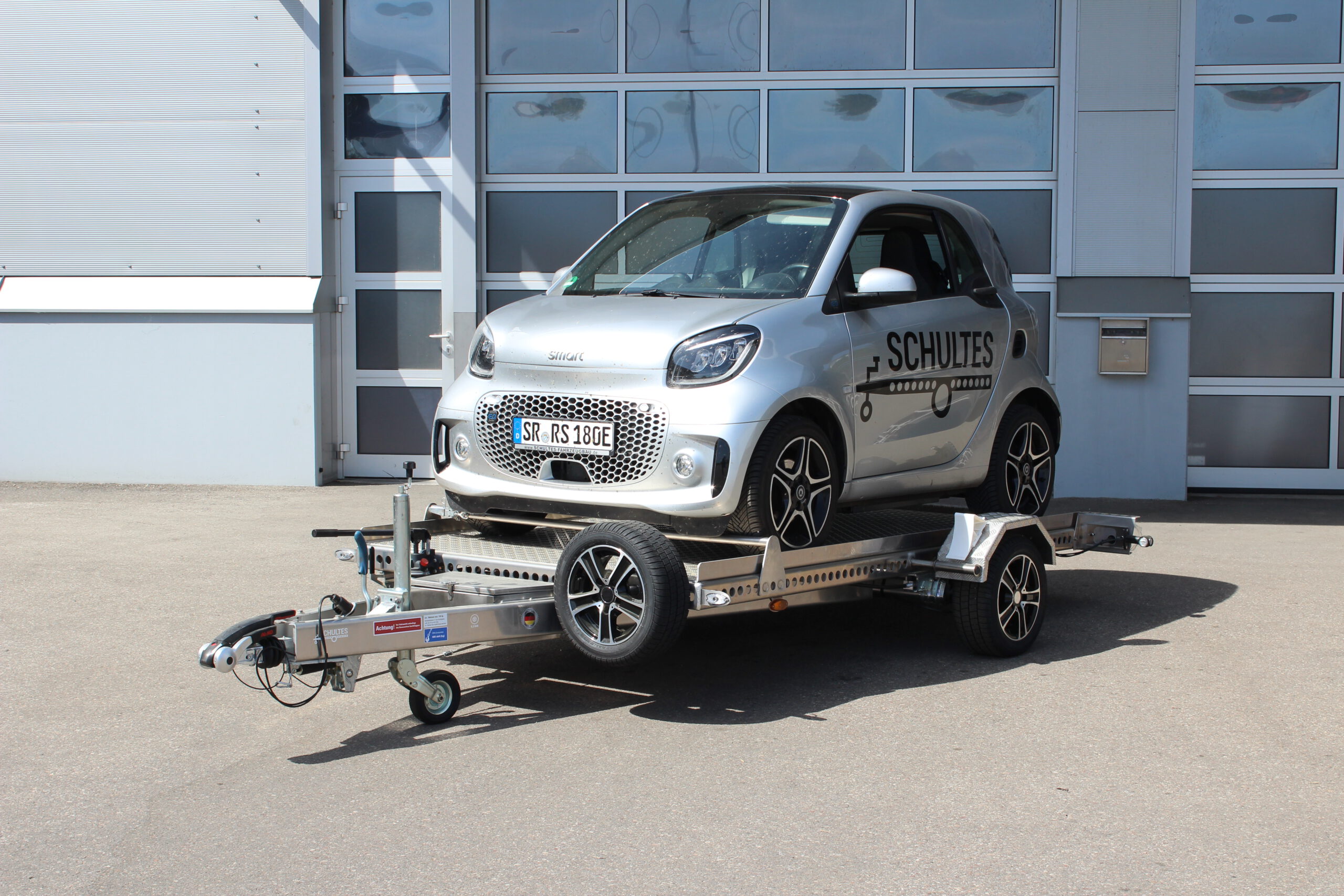 Absenkanhänger aus Aluminium mit einem silbernen Smart fortwo beladen vor einer Halle in einem Hof