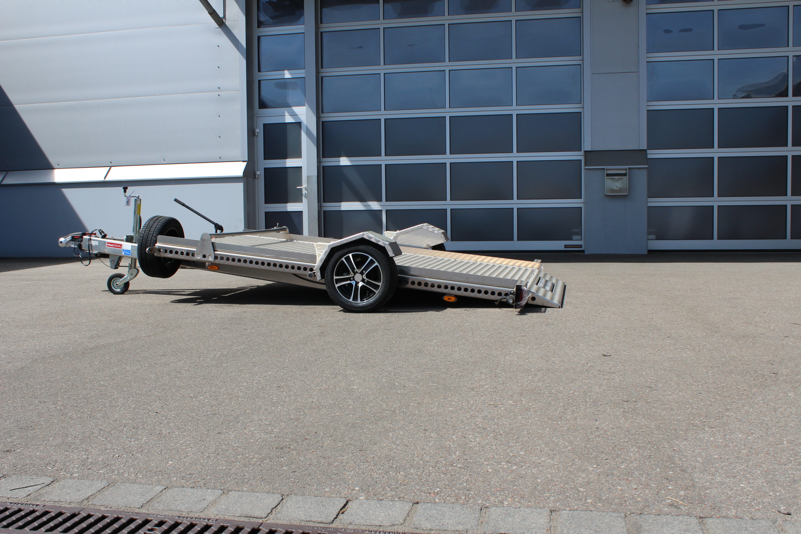 Absenkanhänger aus Aluminium mit einem silbernen Smart fortwo beladen vor einer Halle in einem Hof
