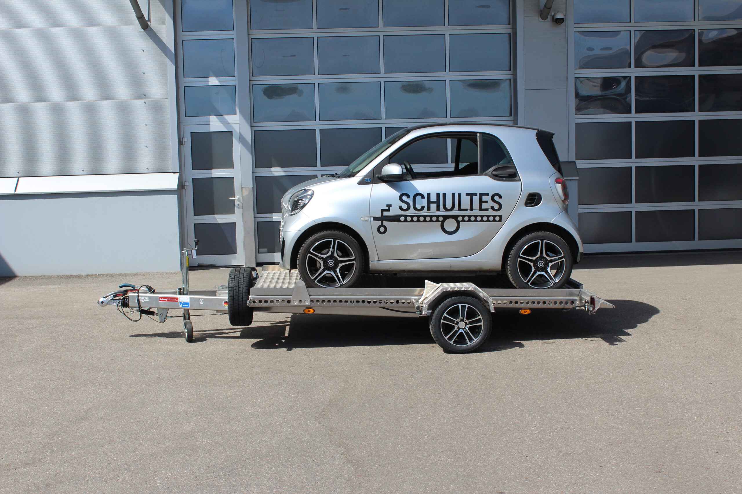 Absenkanhänger aus Aluminium mit einem silbernen Smart fortwo beladen vor einer Halle in einem Hof