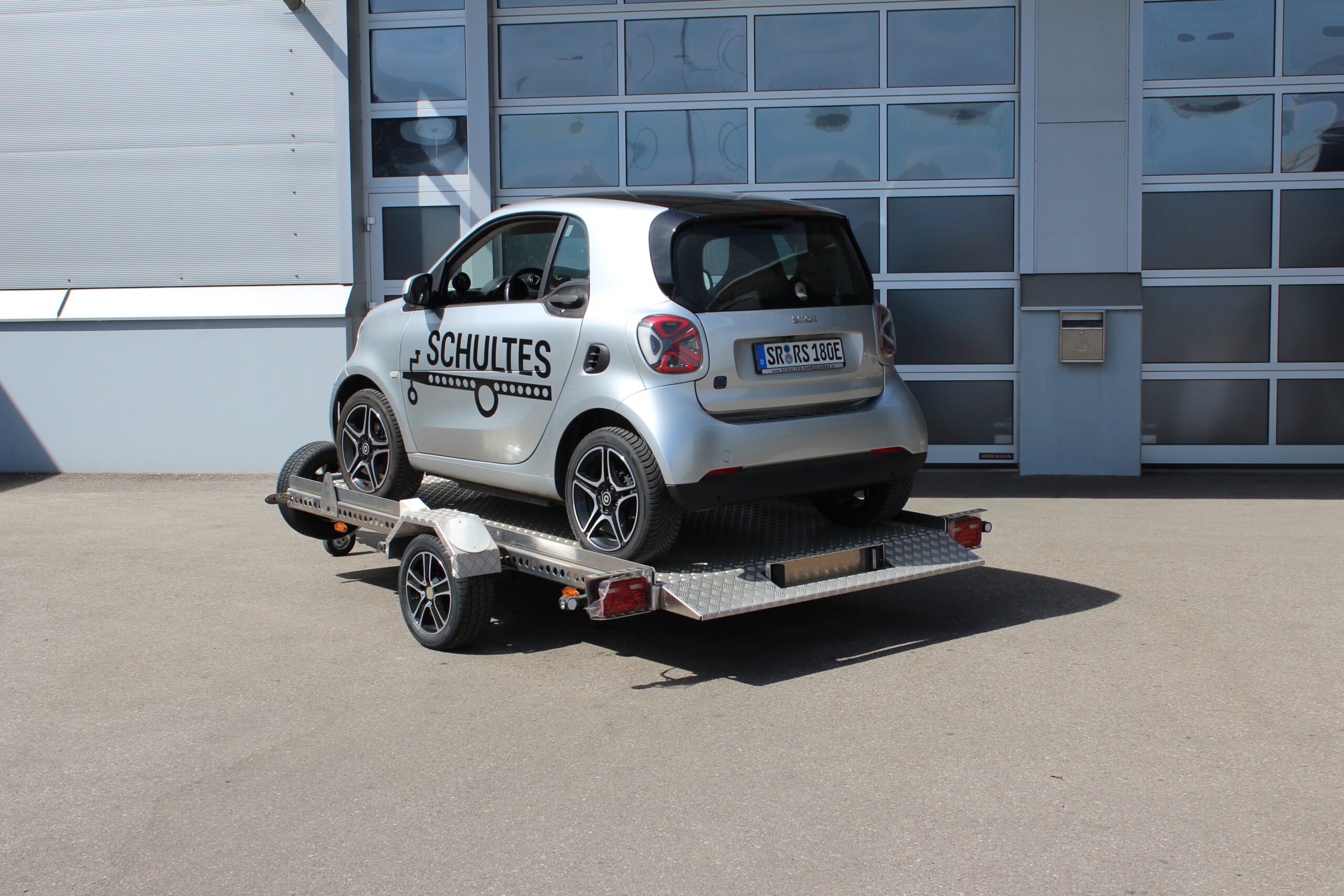 Absenkanhänger aus Aluminium mit einem silbernen Smart fortwo beladen vor einer Halle in einem Hof