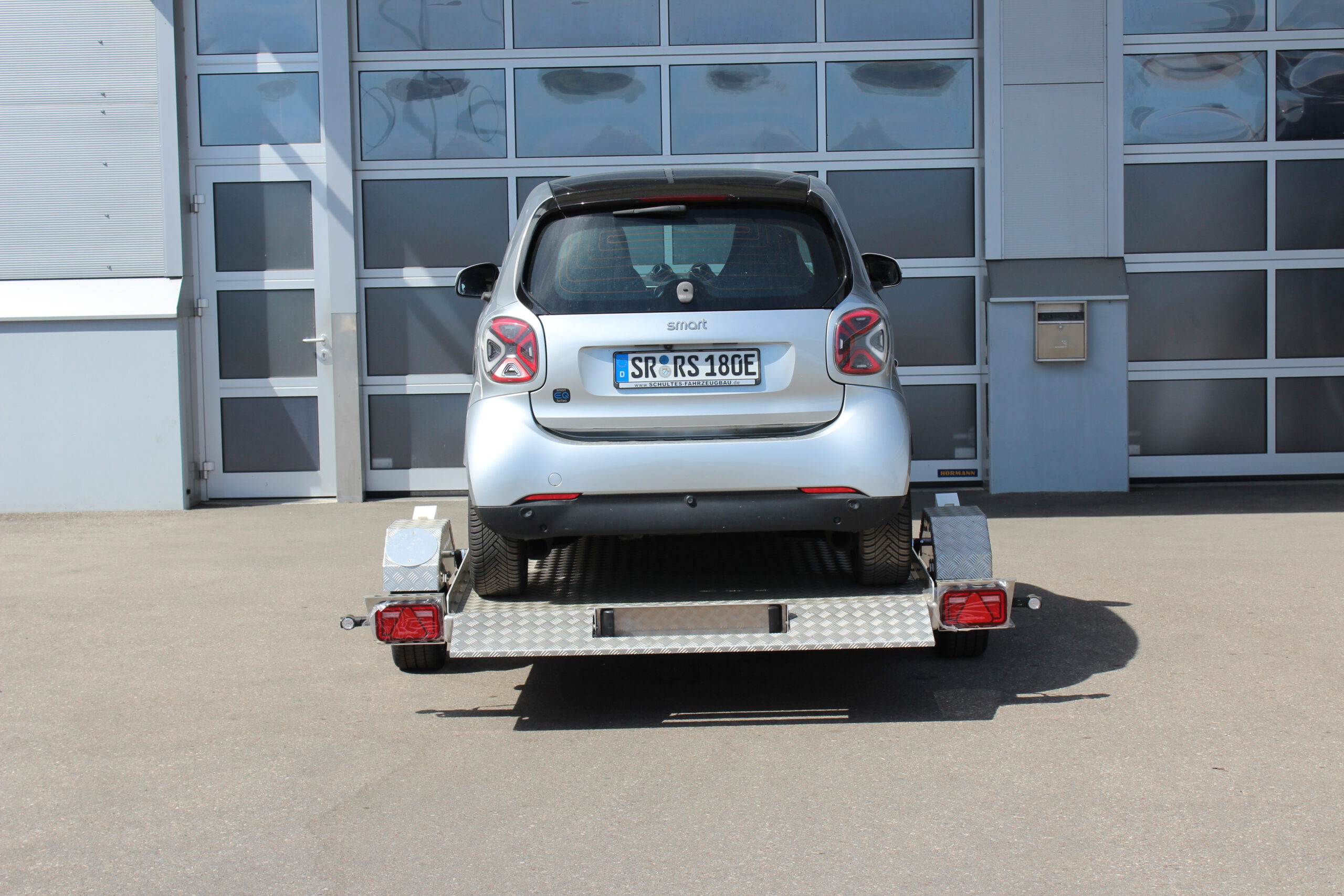 Absenkanhänger aus Aluminium mit einem silbernen Smart fortwo beladen vor einer Halle in einem Hof