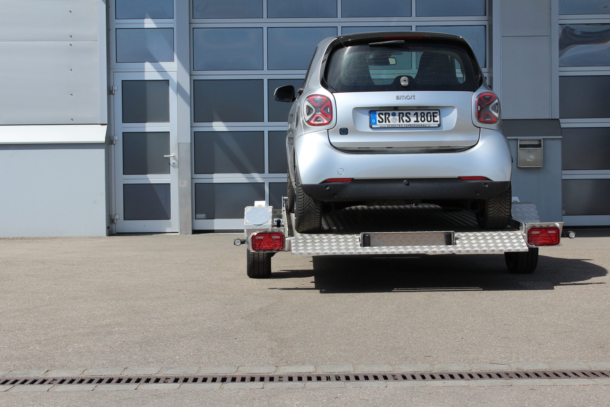 Absenkanhänger aus Aluminium mit einem silbernen Smart fortwo beladen vor einer Halle in einem Hof