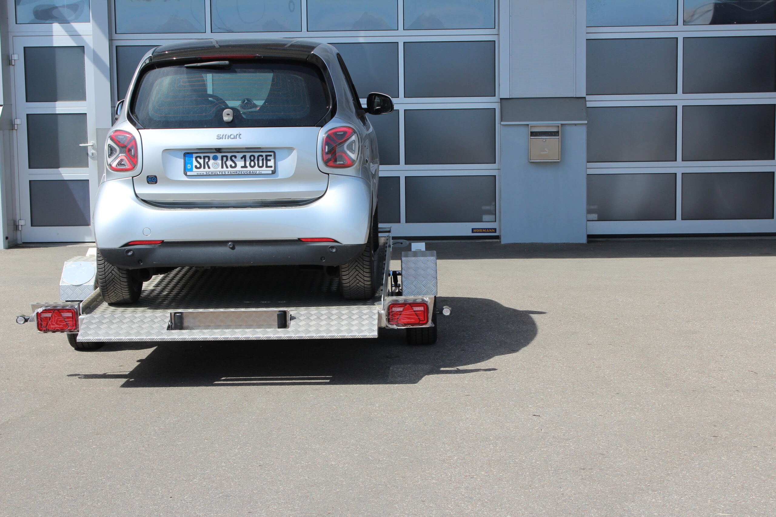 Absenkanhänger aus Aluminium mit einem silbernen Smart fortwo beladen vor einer Halle in einem Hof