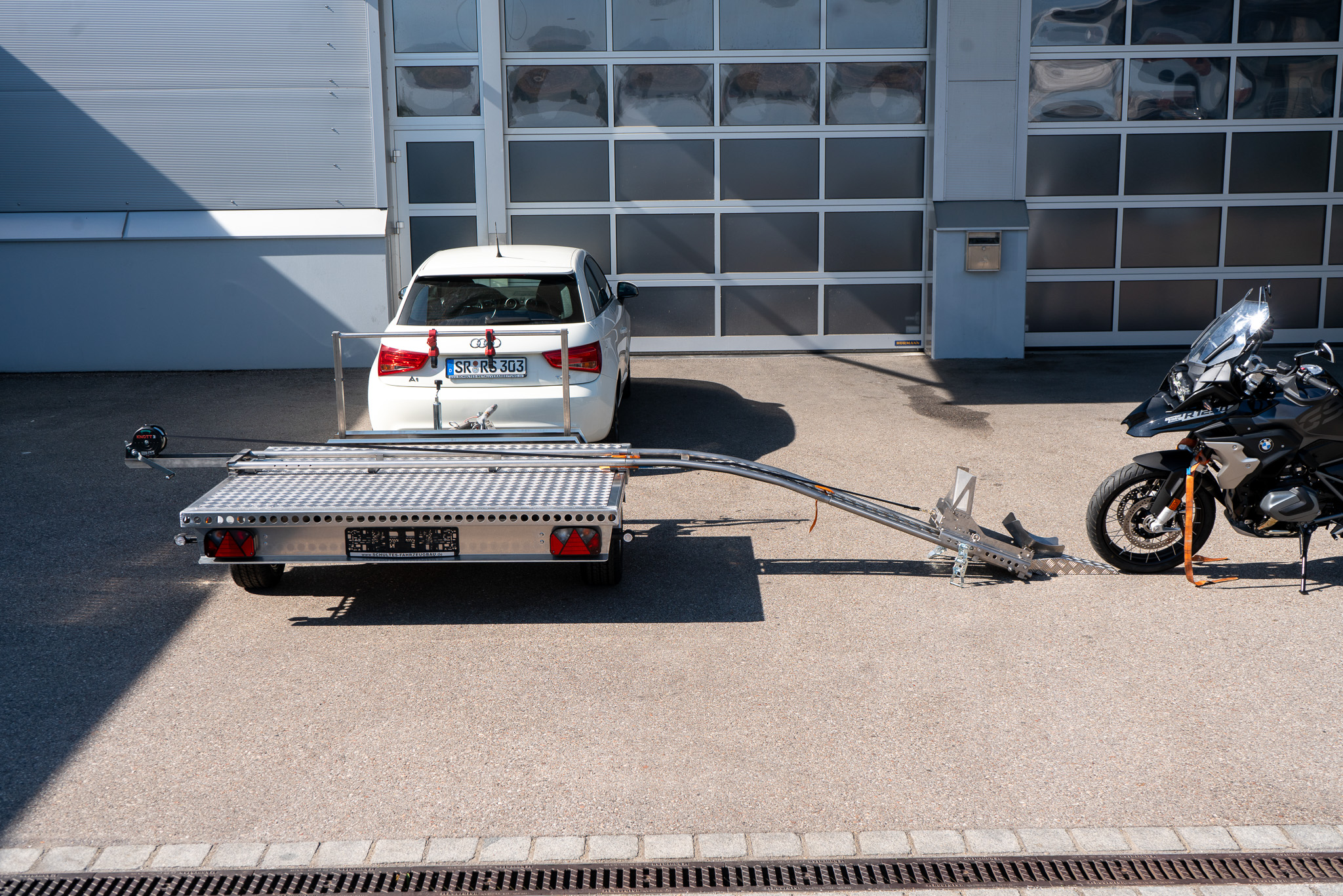 Motorrad Verladesystem auf einem Anhänger befestigt, hinter einem Auto - vor einer Halle - Seitenansicht
