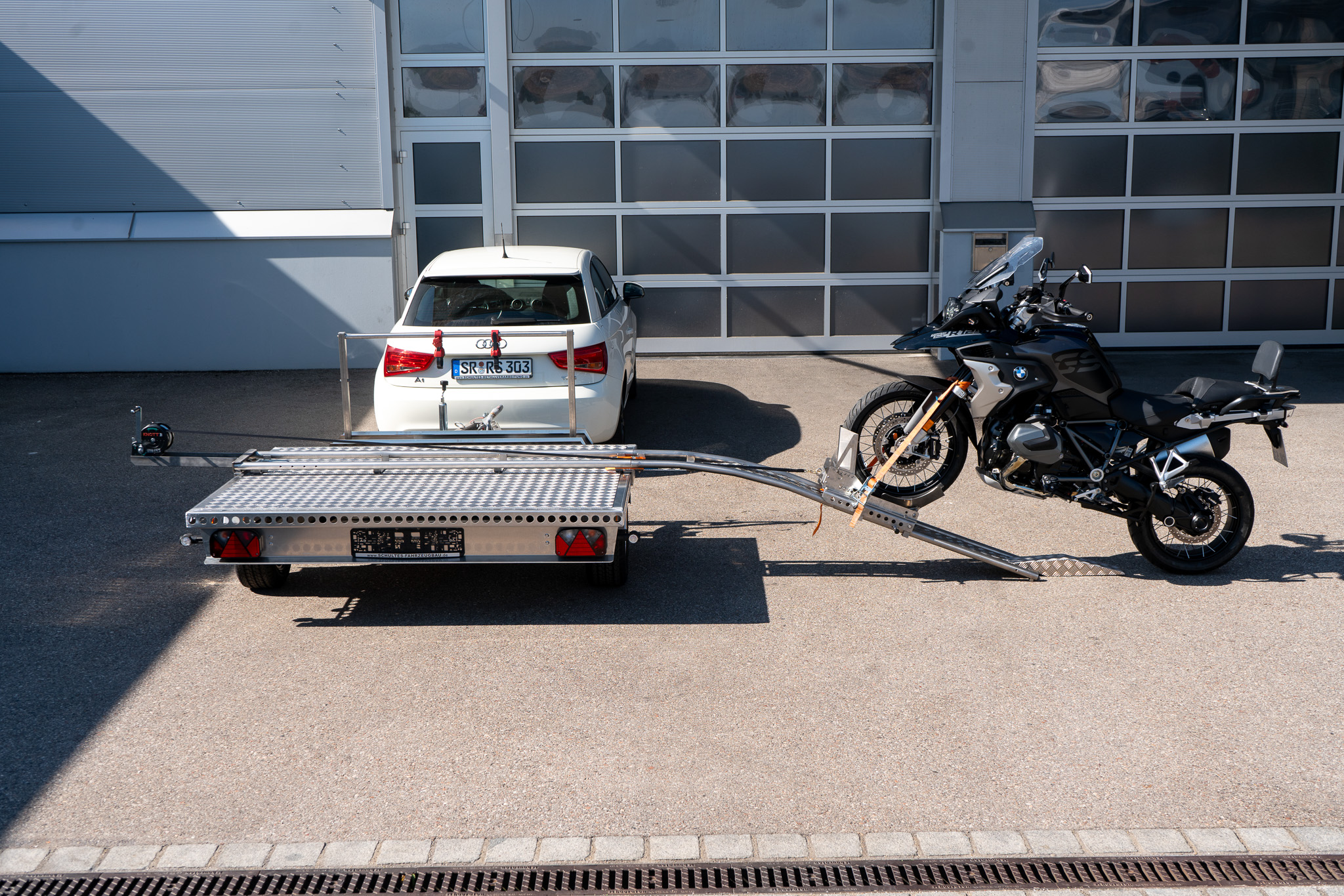 Motorrad Verladesystem auf einem Anhänger befestigt. Motorrad wird auf den Anhänger gezogen - hinter einem Auto - vor einer Halle - Seitenansicht