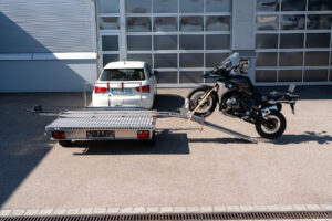 Motorrad Verladesystem auf einem Anhänger befestigt. Motorrad wird auf den Anhänger gezogen - hinter einem Auto - vor einer Halle - Seitenansicht