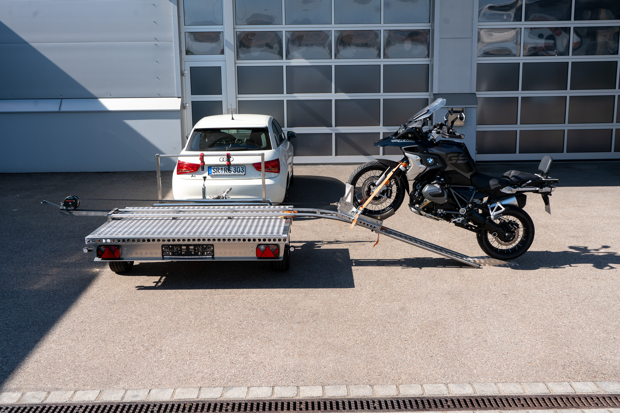 Motorrad Verladesystem auf einem Anhänger befestigt. Motorrad wird auf den Anhänger gezogen - hinter einem Auto - vor einer Halle - Seitenansicht
