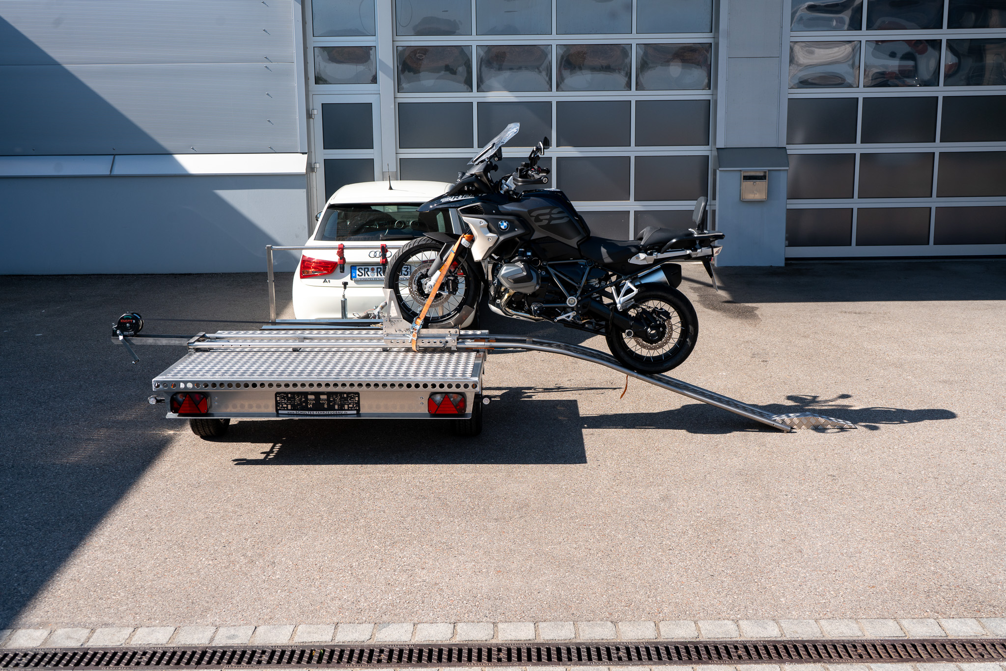 Motorrad Verladesystem auf einem Anhänger befestigt. Motorrad wird auf den Anhänger gezogen - hinter einem Auto - vor einer Halle - Seitenansicht