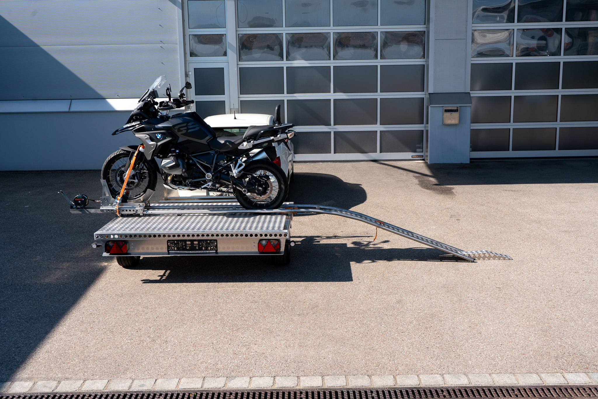 Motorrad Verladesystem auf einem Anhänger befestigt. Motorrad wird auf den Anhänger gezogen - hinter einem Auto - vor einer Halle - Seitenansicht