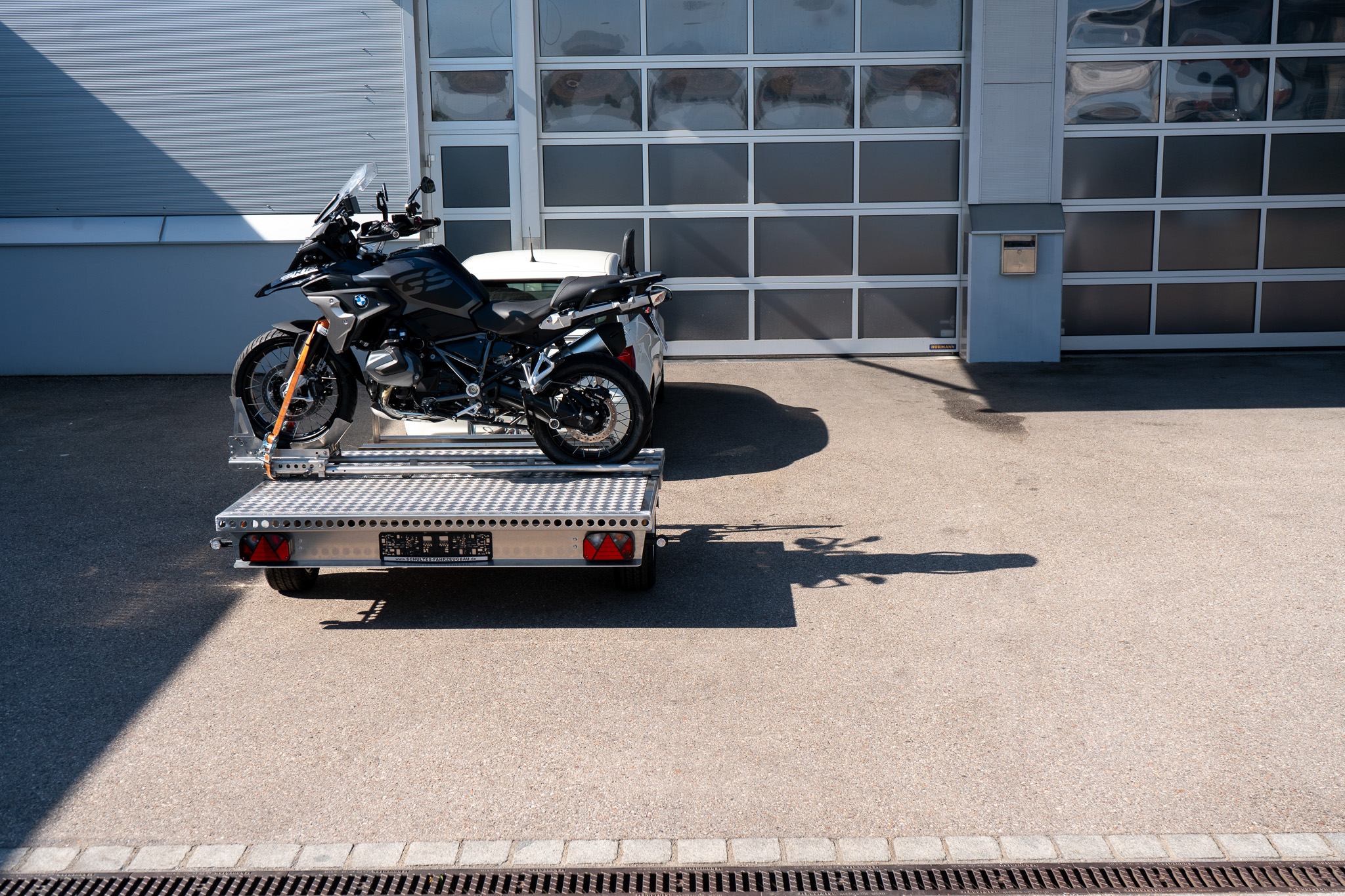 Motorrad Verladesystem auf einem Anhänger befestigt. Motorrad wird auf den Anhänger gezogen - hinter einem Auto - vor einer Halle - Seitenansicht
