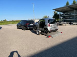 Smart fortwo auf abgesenktem Absenkanhänger hinter einem Auto in einem Hof