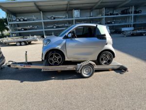 Smart fortwo auf abgesenktem Absenkanhänger hinter einem Auto in einem Hof - von der Seite