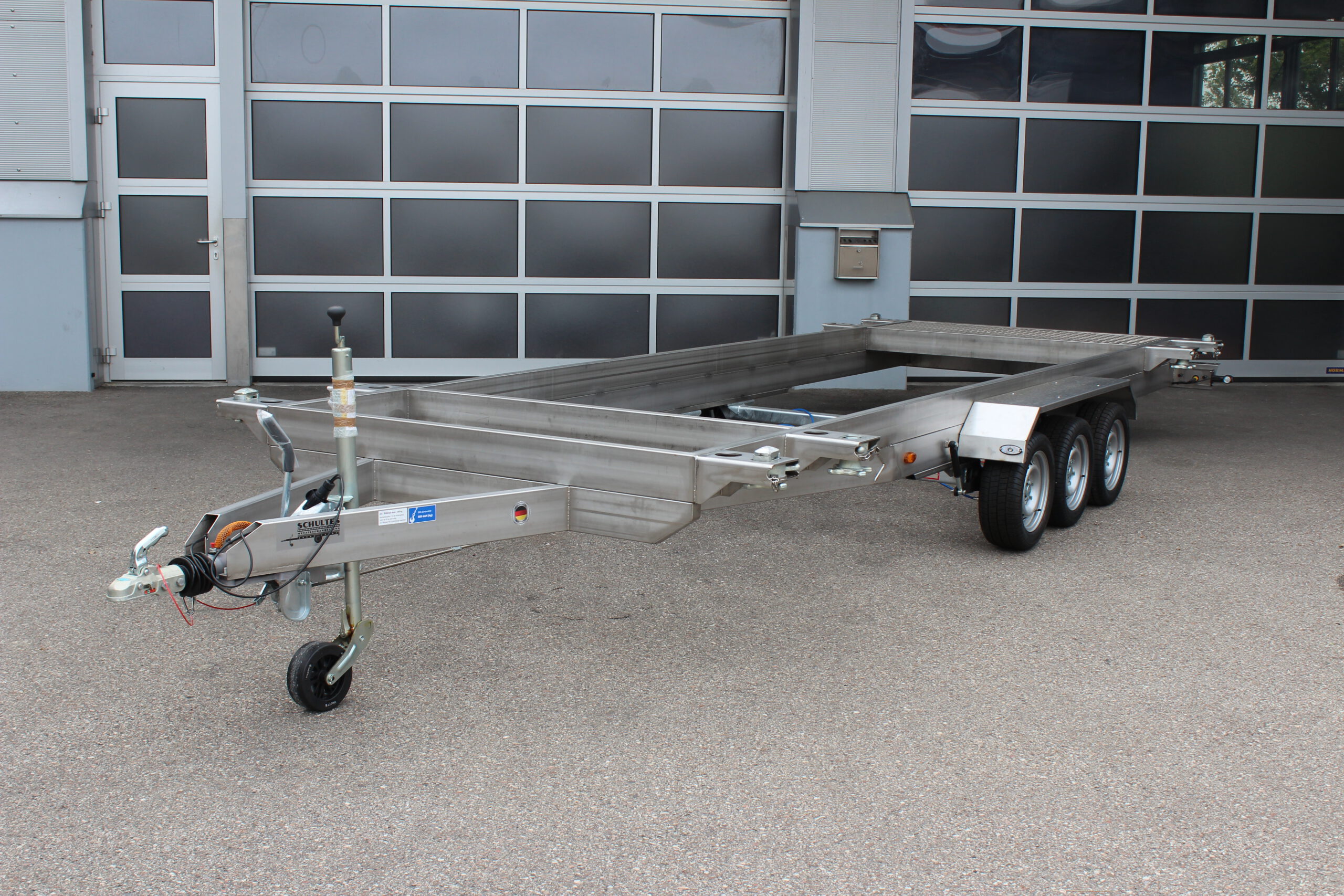 Anhänger aus Edelstahl zum Transport von Überseecontainern. Anhänger steht vor einem Hallentor. Anhängerfahrgestell ohne Bodenplatte für Containertransport.