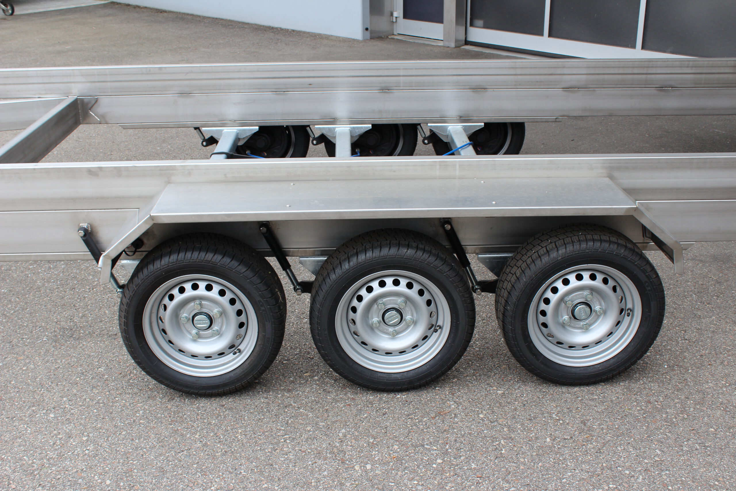 Anhänger aus Edelstahl zum Transport von Überseecontainern. Anhänger steht vor einem Hallentor. Anhängerfahrgestell ohne Bodenplatte für Containertransport.