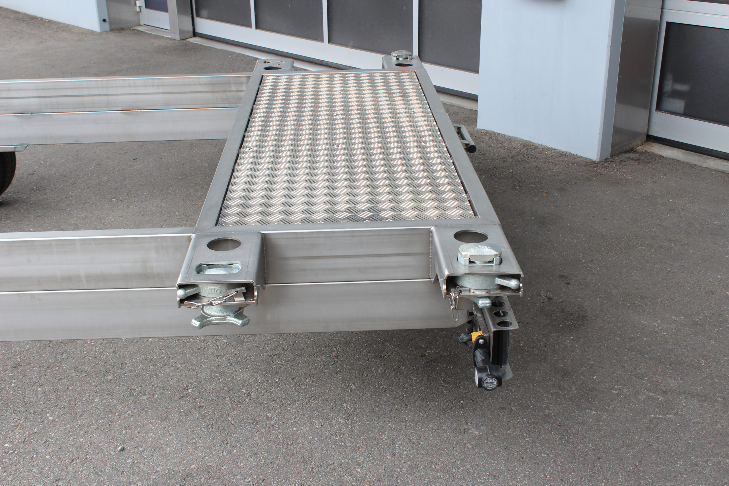Anhänger aus Edelstahl zum Transport von Überseecontainern. Anhänger steht vor einem Hallentor. Anhängerfahrgestell ohne Bodenplatte für Containertransport.