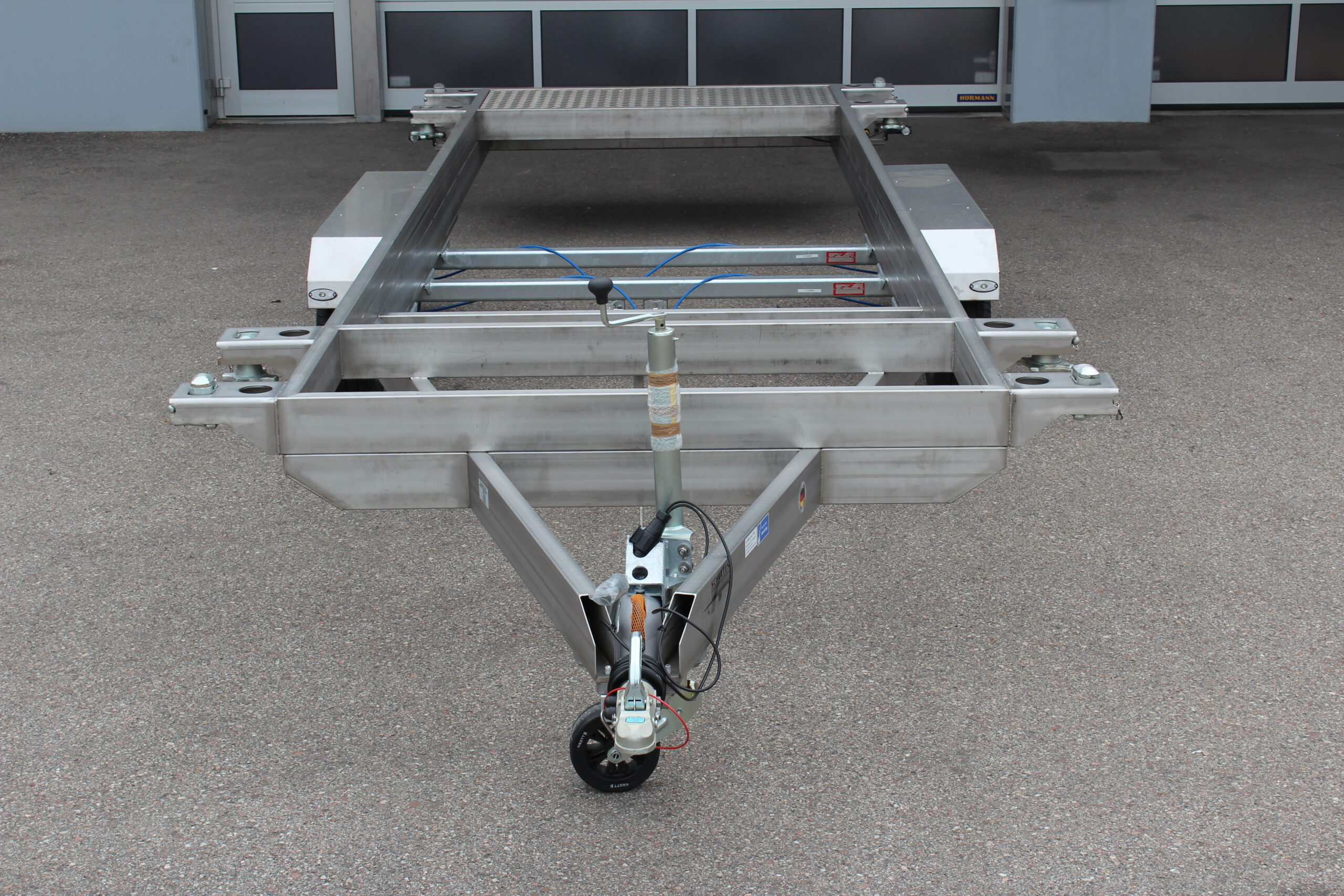 Anhänger aus Edelstahl zum Transport von Überseecontainern. Anhänger steht vor einem Hallentor. Anhängerfahrgestell ohne Bodenplatte für Containertransport.