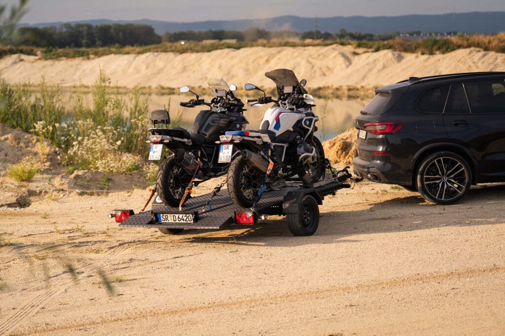 Schwarzer Absenkanhänger mit zwei BMW GS Motorrädern beladen an einem BMW X5 in einer Kiesgrube