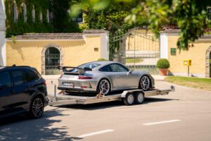 Edelstahl Tieflader Anhänger mit einem Porsche GT3 beladen vor einem gelben Haus