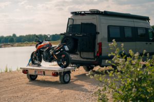Kleiner Anhänger mit einem Motorrad beladen an einem Sprinter vor einem See