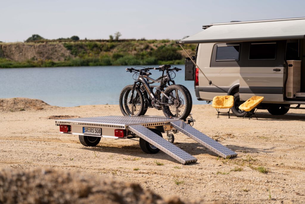 Kleiner Plattformanhänger mit Zwei Fahrrädern beladen vor einem Sprinter an einem See