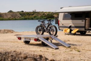 Kleiner Plattformanhänger mit Zwei Fahrrädern beladen vor einem Sprinter an einem See
