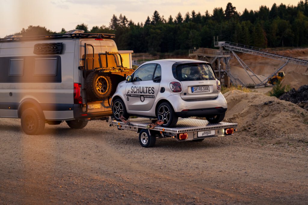 Anhänger für Autos mit einem Smart beladen hinter einem Wohnmobil vor einer Kiesgrube