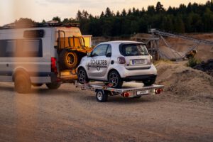 Anhänger für Autos mit einem Smart beladen hinter einem Wohnmobil vor einer Kiesgrube