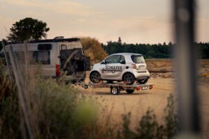 Anhänger für Autos mit einem Smart beladen hinter einem Wohnmobil vor einer Kiesgrube