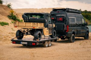 schwarzer Plattformanhänger mit einem sandfarbenen UTV beladen hinter einem schwarzen Sprinter vor einem See