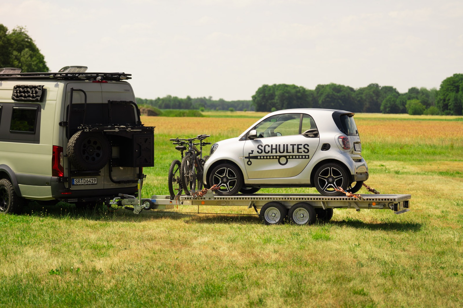 UA350 Tandem Anhänger Smart und Fahrräder (2) Anhänger aus Aluminium mit Smart fortwo und zwei Fahrrädern hinter einem Mercedes Sprinter vor Bäumen und einer grünen Wiese
